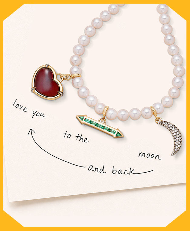 A pearl necklace with three 18k gold charms: a red heart pendant, a green arrow pendant, and a crescent moon pendant, displayed on a cream card with handwritten text reading 'love you to the moon and back' inside a yellow border.